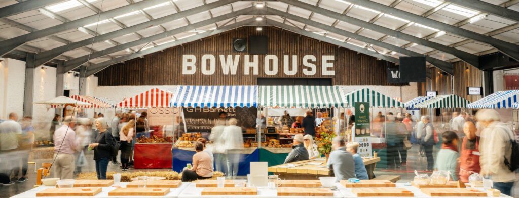 A lively local and organic food market in a big hanger with the words Bowhouse displayed above the traders